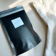 Load image into Gallery viewer, Vintage Alchemy Coconut Milk Bath in matte black resealable pouch with soft linen and warm neutral stone surface styling