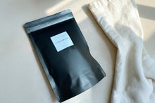 Load image into Gallery viewer, Vintage Alchemy Coconut Milk Bath in matte black resealable pouch with soft linen and warm neutral stone surface styling