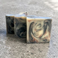 Load image into Gallery viewer, Small-batch Marlowe cold process soap with charcoal, cream, and blackberry marbled swirls displayed on a textured stone surface.