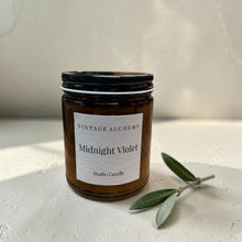 Load image into Gallery viewer, Midnight Violet studio candle in amber glass jar with olive branch on soft plaster background with warm natural light