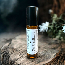 Load image into Gallery viewer, Roll-on bottle of 'Vintage Alchemy Orchid Moon' on a wooden surface with flowers in the background
