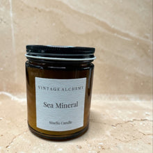 Load image into Gallery viewer, Sea Mineral scented candle in amber glass jar with black lid on warm beige stone background
