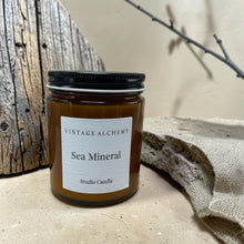 Load image into Gallery viewer, Sea Mineral studio candle in amber glass jar with black lid, weathered wood, linen fabric, and soft botanical shadows on warm stone surface background