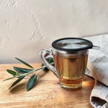 Load image into Gallery viewer, Stainless steel loose leaf tea infuser with lid brewing tea in a glass mug on a warm wood surface with linen and olive branch