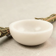 Load image into Gallery viewer, White marble smudge bowl displayed on a neutral stone surface with bundled cleansing herbs in the background.