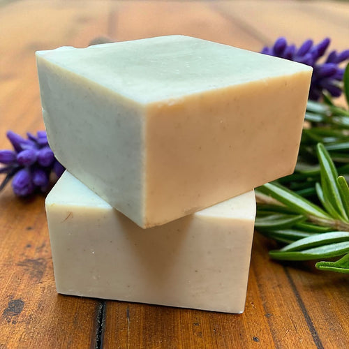 Two stacked soap bars on a wooden surface with lavender and rosemary leaves.