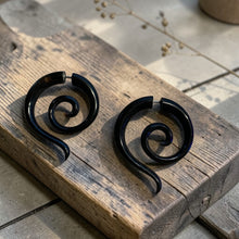 Load image into Gallery viewer, Black spiral fake gauge earrings displayed on weathered wood block with soft botanical background in natural light
