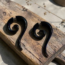 Load image into Gallery viewer, Black spiral fake gauge tribal earrings displayed on weathered wood block with soft botanical background in natural light

