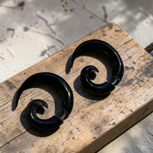 Load image into Gallery viewer, Black spiral tribal fake gauge earrings displayed on weathered wood board with soft botanical background in natural light
