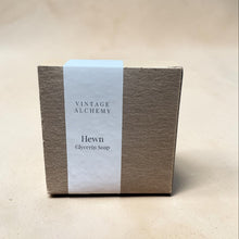 Load image into Gallery viewer, Minimal kraft box packaging for Hewn glycerin soap with a white wrap label on a seamless warm neutral plaster surface.
