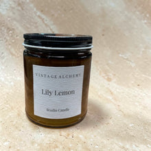 Load image into Gallery viewer, Lily Lemon scented candle in amber glass jar with black lid on warm beige stone background

