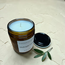 Load image into Gallery viewer, Open Midnight Violet studio candle in amber glass jar with black lid and olive branch on soft plaster background
