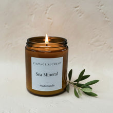 Load image into Gallery viewer, Lit Sea Mineral studio candle in amber glass jar with minimalist white label on warm textured plaster surface background

