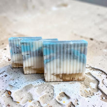 Load image into Gallery viewer, Three ribbed glycerin soap bars in layered coastal tones of blue, gray, cream, and sand displayed on a textured shoreline-inspired surface.
