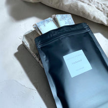 Load image into Gallery viewer, Matte black wax melt refill pouch with botanical soy wax melt bars partially visible on neutral plaster surface for Vintage Alchemy Uncharted fragrance.
