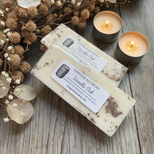 Two wax melts with 'Vintage Alchemy' labels and two lit candles on a wood surface.